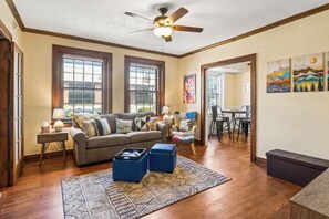 A living room with a sofa, ottomans, a patterned rug, a ceiling fan, and large windows. - The Flat on Fifth (Knoxville)