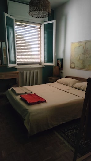 A bedroom with a bed, a window with blinds, a radiator, a map on the wall, and a small table with a lamp. - New Listing (Ravenna)