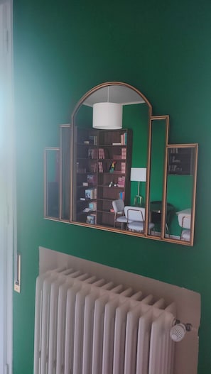 Una habitación con pared verde, un radiador, una estantería y un espejo con marco de madera.