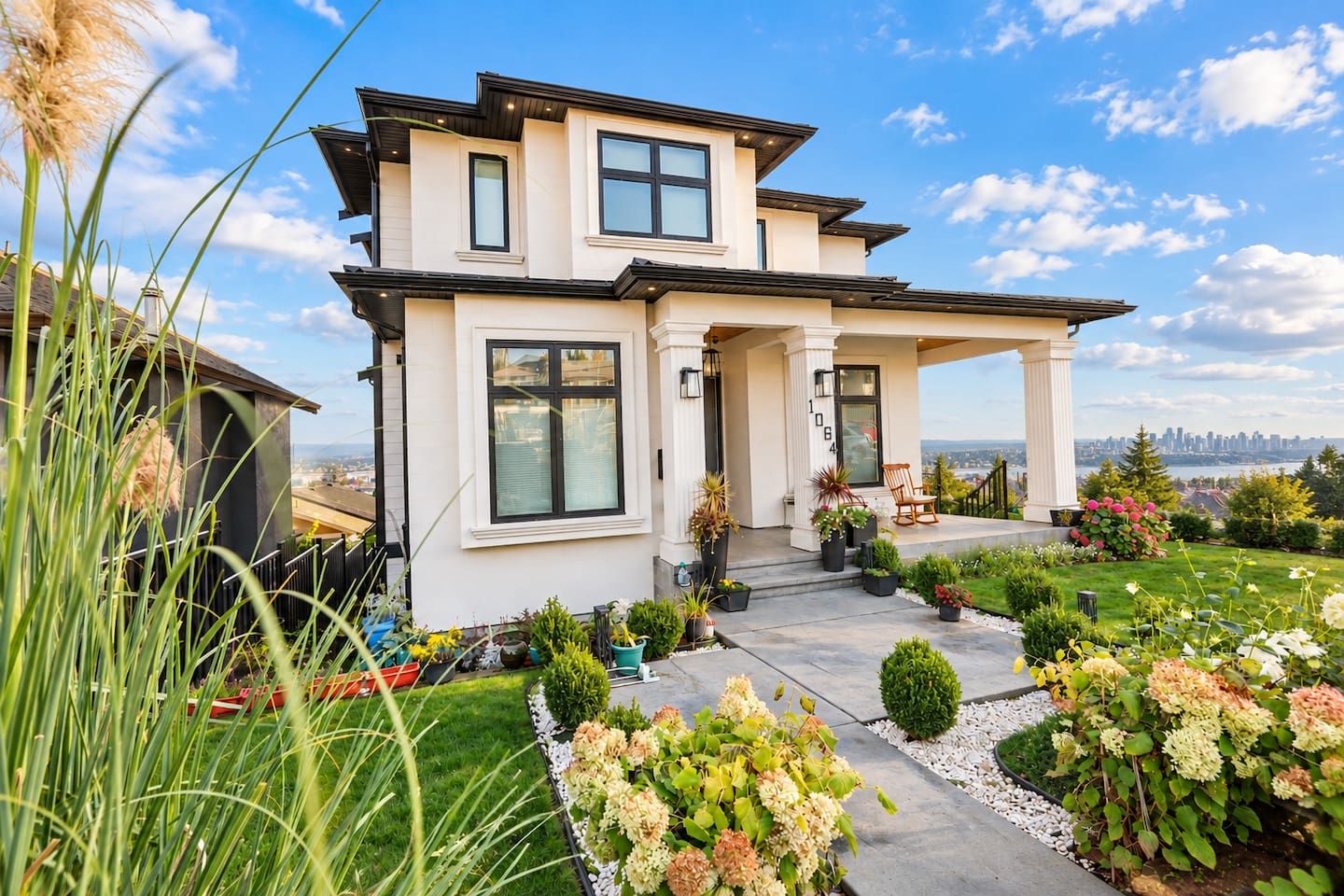 A modern two-story house with a covered porch, a view of the city skyline, and a well-maintained garden with various plants and flowers.