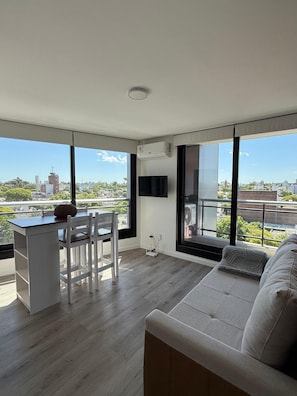 A modern living space with a sofa, dining table, and chairs, large windows with a city view, and an air conditioning unit. - Espectacular loft en Buceo con amenities de lujo (Montevideo)