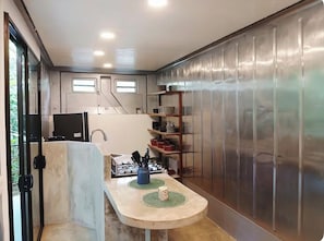 Eine kompakte Küche mit Gaskochfeld, Spüle und einer kleinen Arbeitsfläche. Zwei Fenster sorgen für natürliches Licht, und eine Wandverkleidung aus Metall ist an einer Seite angebracht.