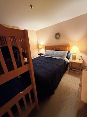 A bedroom with a bunk bed, a single bed, two bedside tables with lamps, a wooden headboard, and a wall-mounted light fixture.