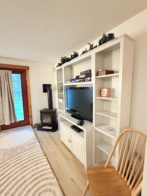 A room with a television on a white entertainment unit, a fireplace, a wooden chair, and a window with curtains.