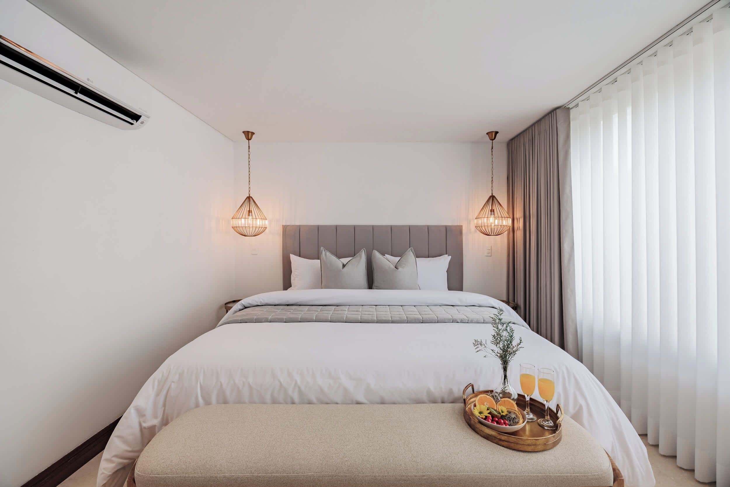 Una habitación de hotel moderna con una cama grande, una mesita de noche con una bandeja de frutas y bebidas, y vistas de la ciudad a través de cortinas translúcidas.