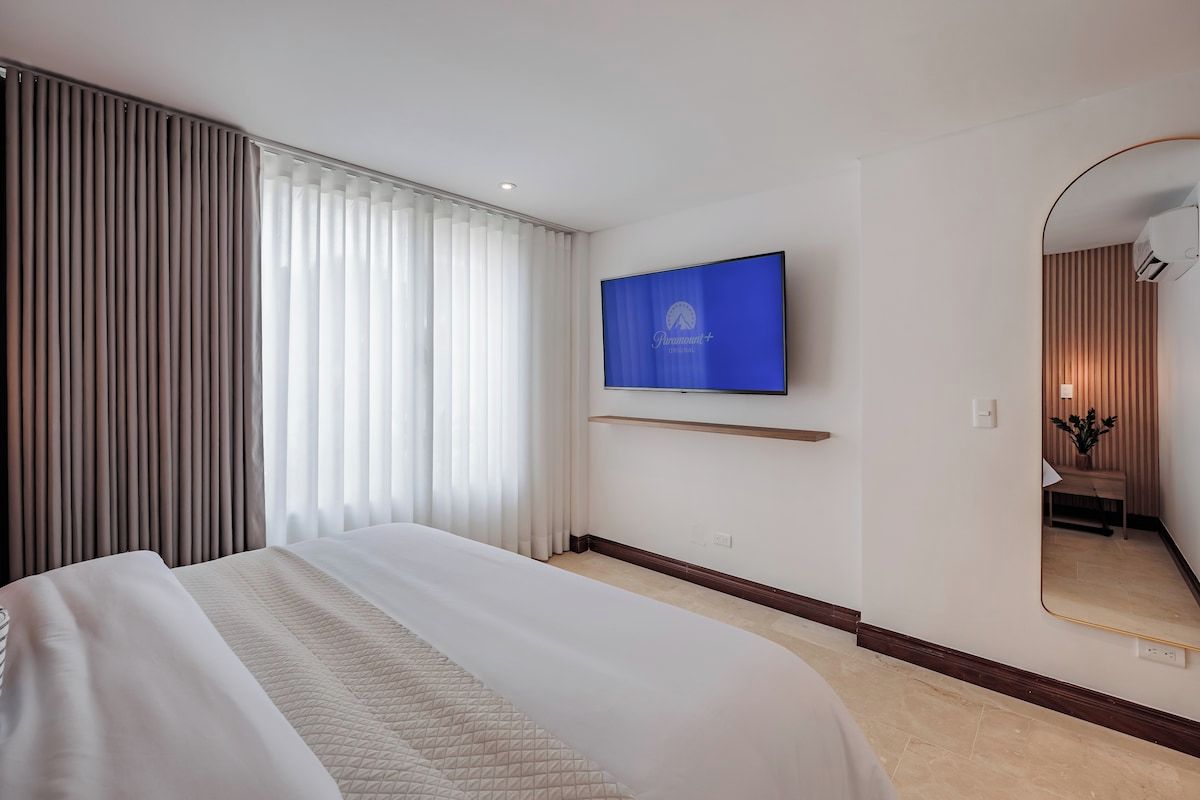 A hotel room with a bed, a flat-screen TV mounted on the wall, a large mirror, and a window with curtains.