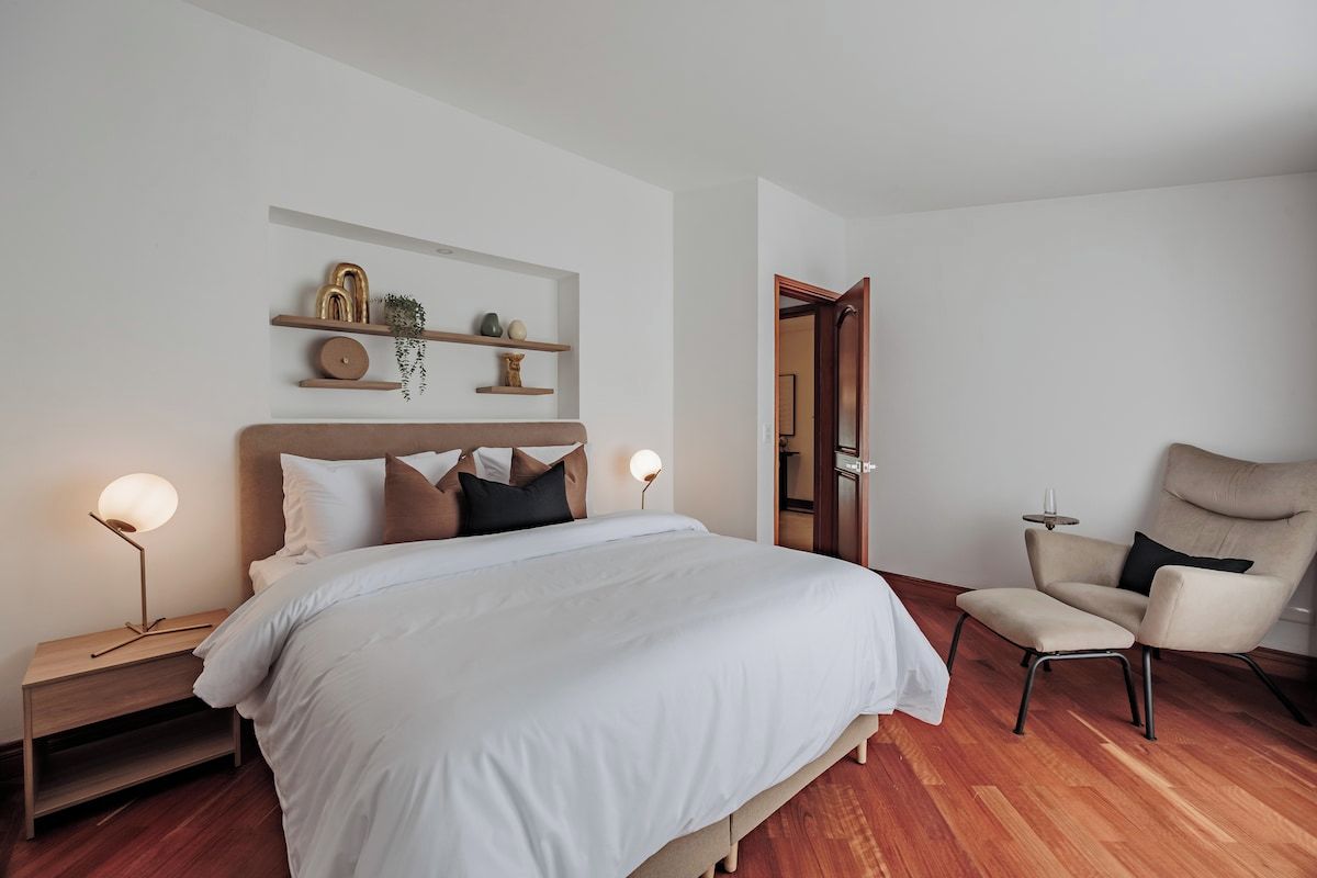 Een slaapkamer met een bed, nachtkastjes, een stoel en een plank met decoratieve objecten.