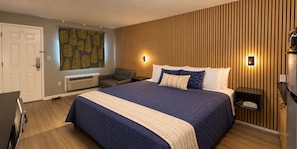 A hotel room with a large bed, a desk, a chair, a window with blinds, and a wall-mounted heater.