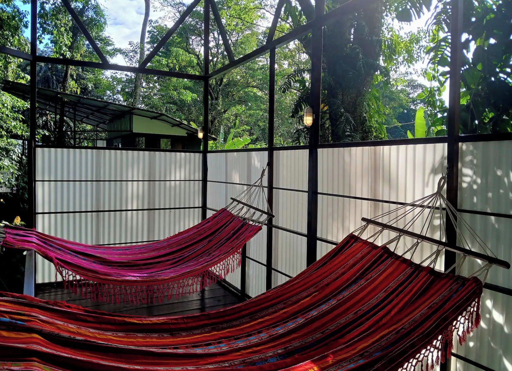 A hammock suspended between metal beams in a covered outdoor area with a view of greenery.