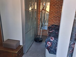 A narrow room with a bed, a suitcase, a box, and a mirror reflecting a brick wall and a window. - New Listing (Madrid)