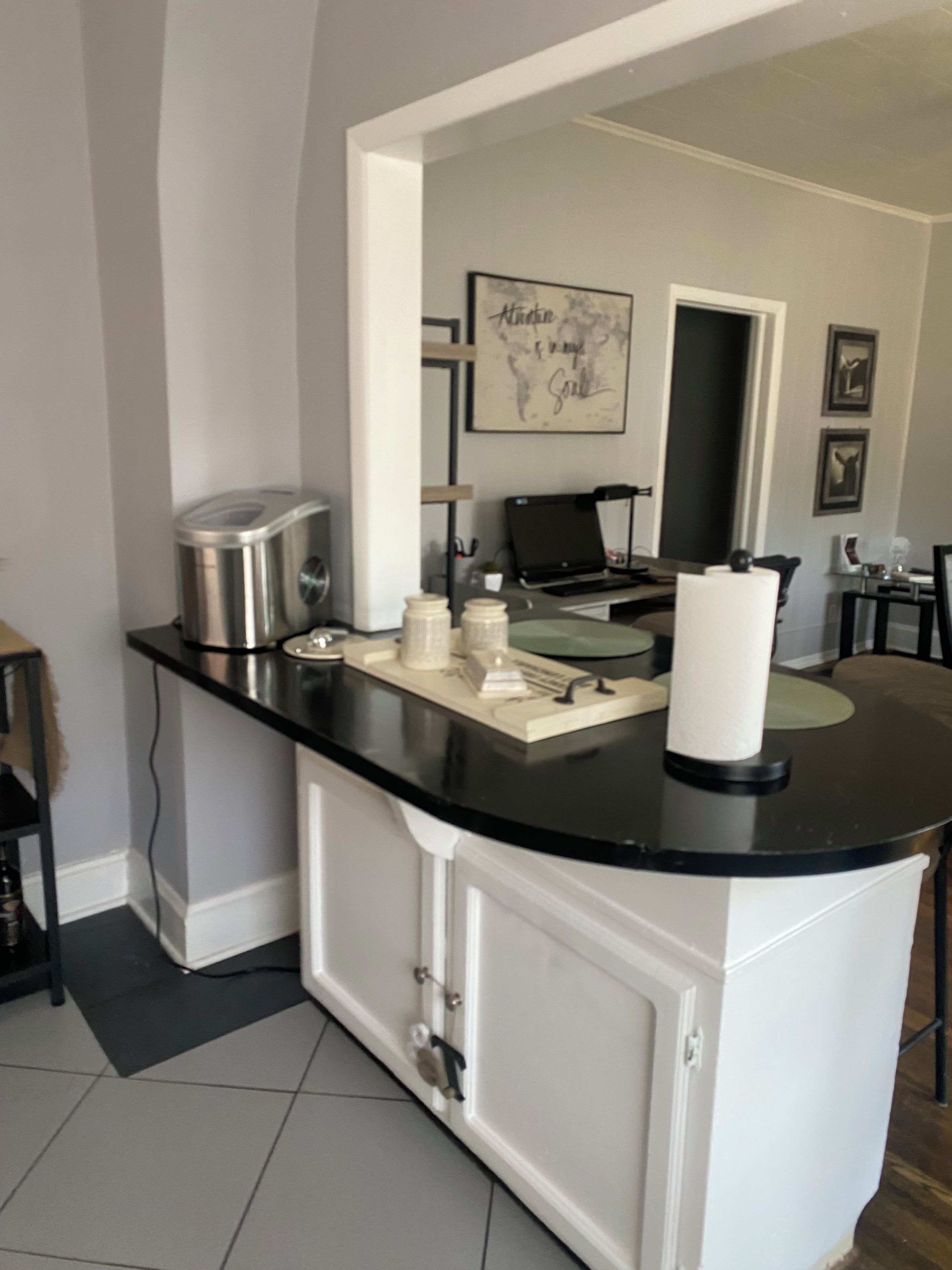 A kitchen with a black countertop, white cabinets, a stainless steel kettle, a coffee maker, and a framed artwork on the wall.
