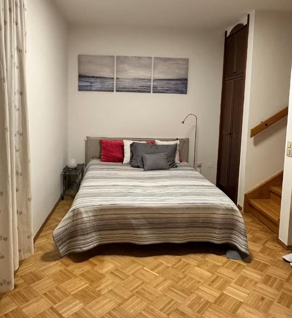 A bedroom with a bed, a nightstand, a lamp, a door, and a staircase.