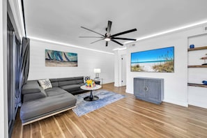 A modern living room with a gray sectional sofa, a small round table with a lamp, a ceiling fan, and artwork on the walls. - Simply Serenity-Lakefront-Pet Friendly-Heated Pool (Destin)
