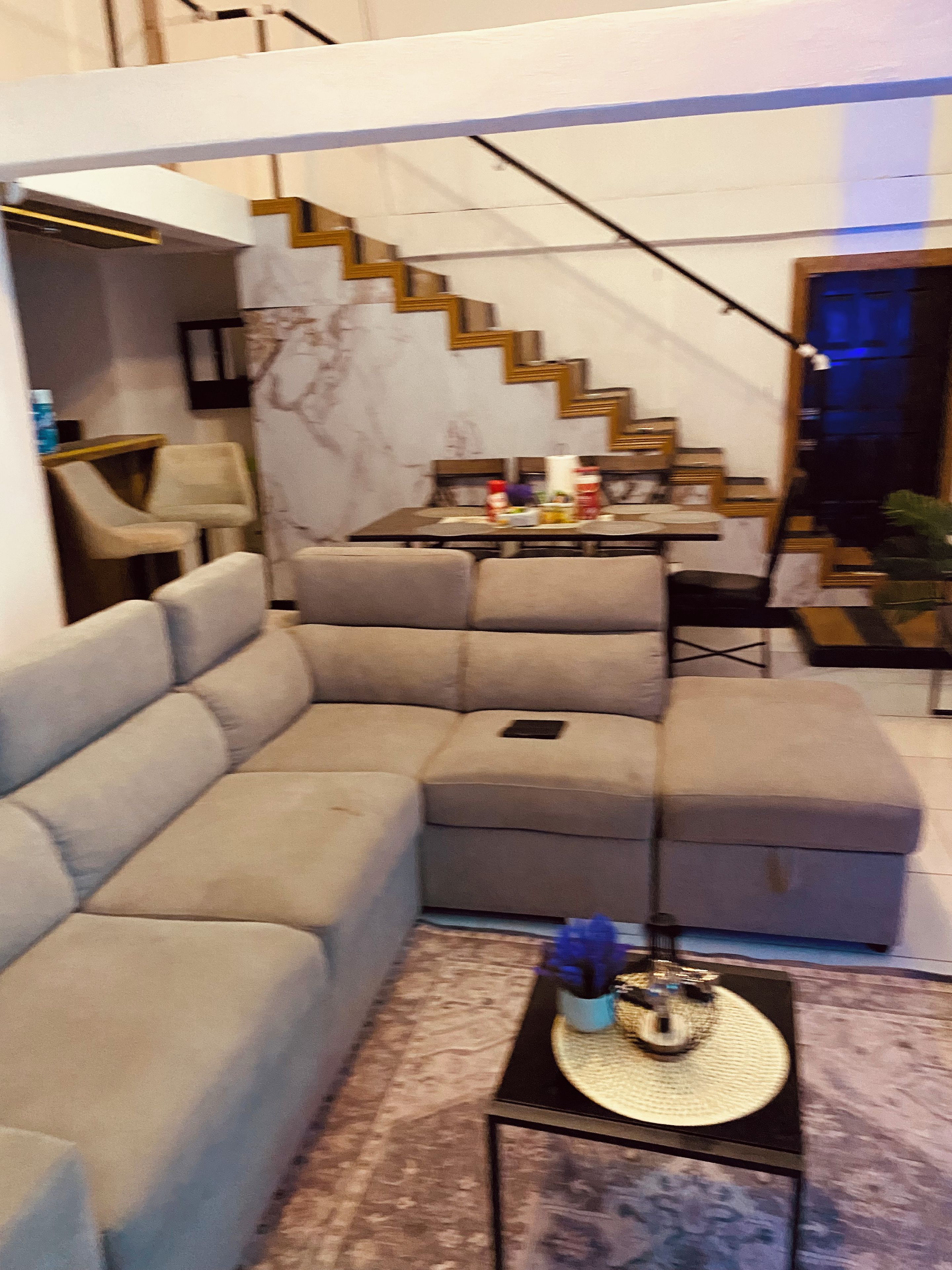 A modern living room with a sectional sofa, a dining table with chairs, and a staircase.