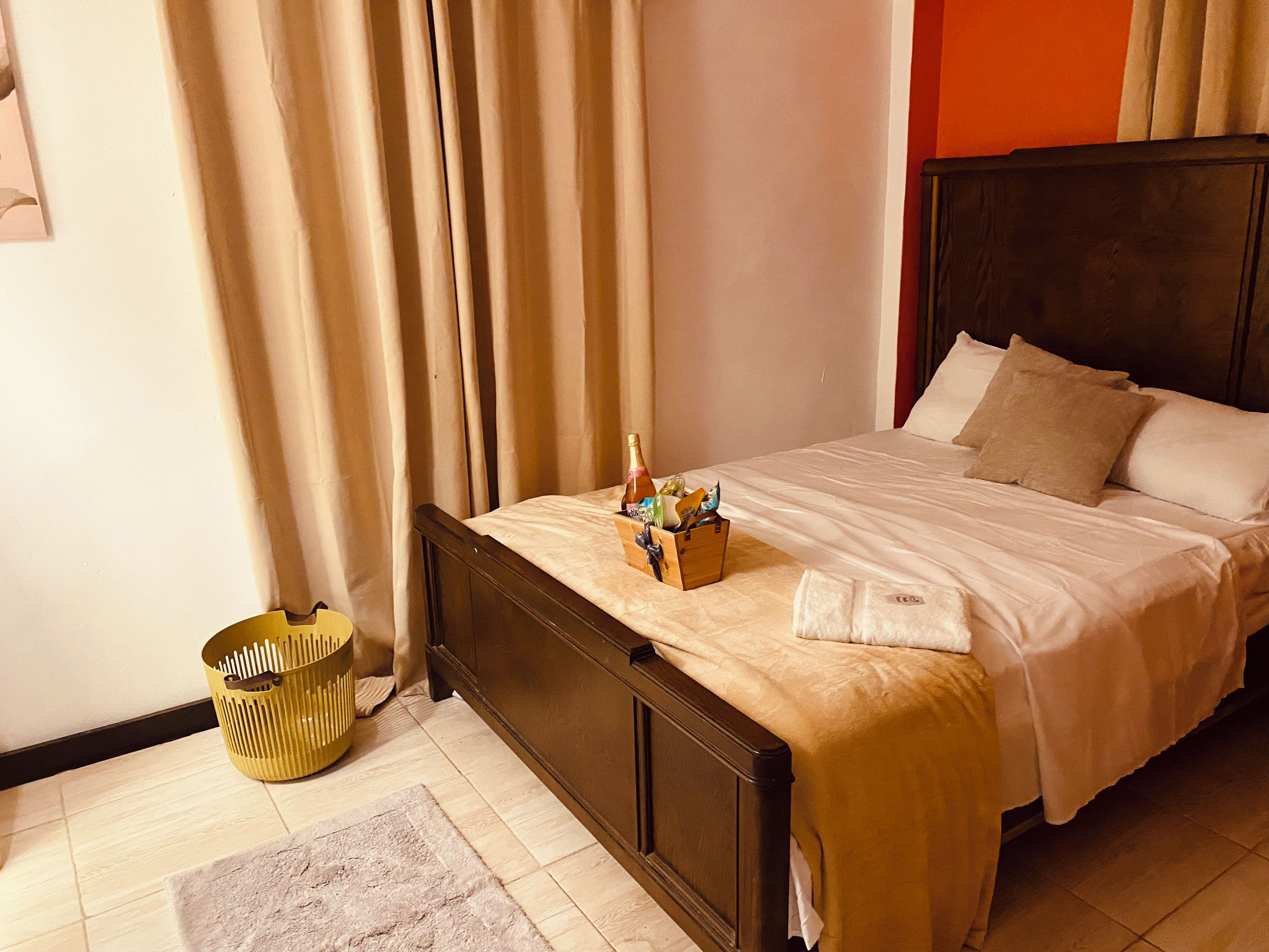 A hotel room with a bed, curtains, a wooden headboard, a bedside table with a bottle and glasses, and a small basket.