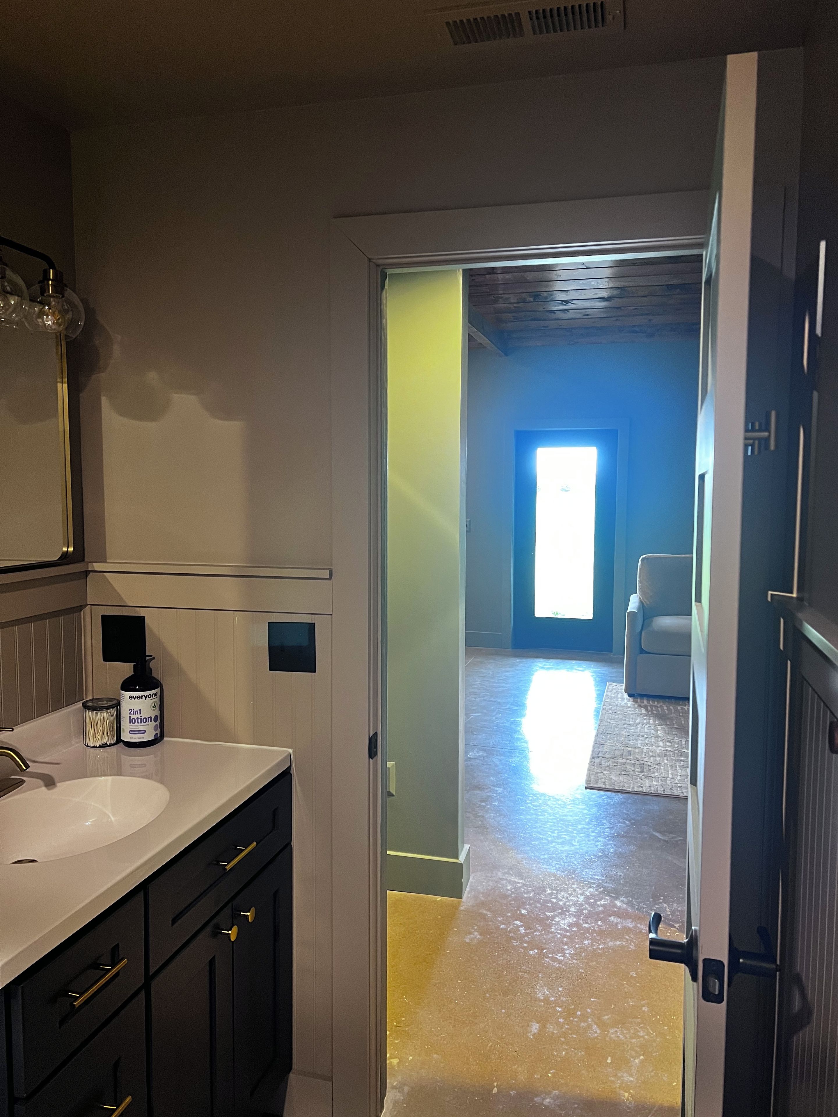 Ein Badezimmer mit Waschbecken, Spiegel und Toilette. Ein Flur führt zu einem Raum mit Stuhl und Fenster.