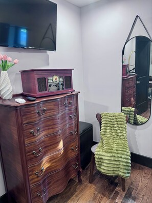 Ein Zimmer mit einem Holzkoffer, einem flachbildfernseher an der Wand, einem Spiegel und einem grünen Handtuch, das über einen Stuhl gelegt ist.