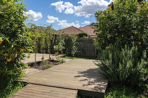 Une terrasse en bois mène à une maison dont le toit est recouvert de tuiles rouges, le tout étant entouré de verdure et d’une clôture en bois.