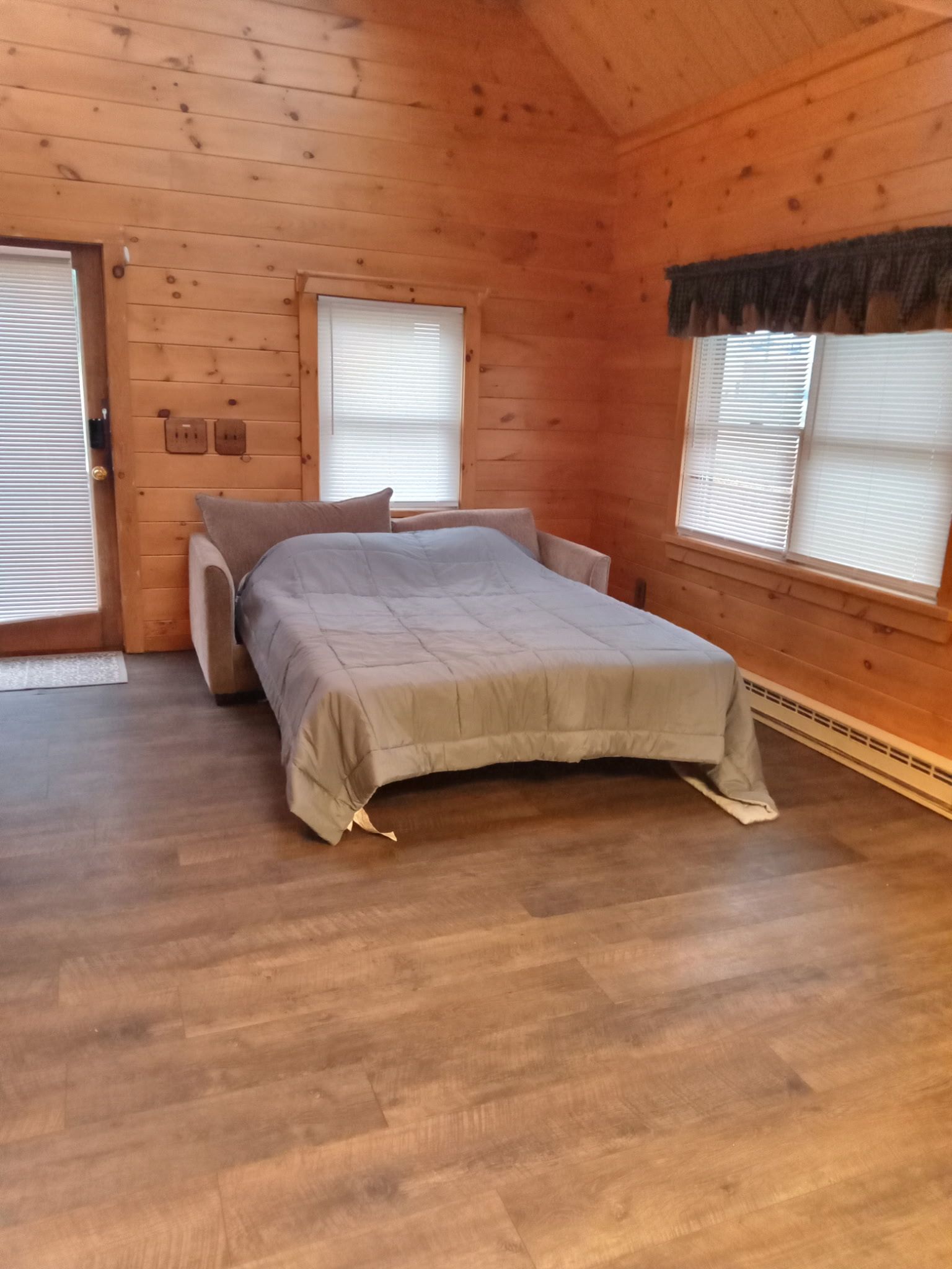 Une chambre dans un chalet en bois, avec un lit, deux fenêtres équipées de stores et une porte.