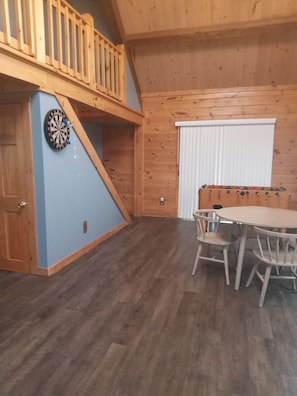 A room with a wooden table and chairs, a dartboard, and a window with blinds.