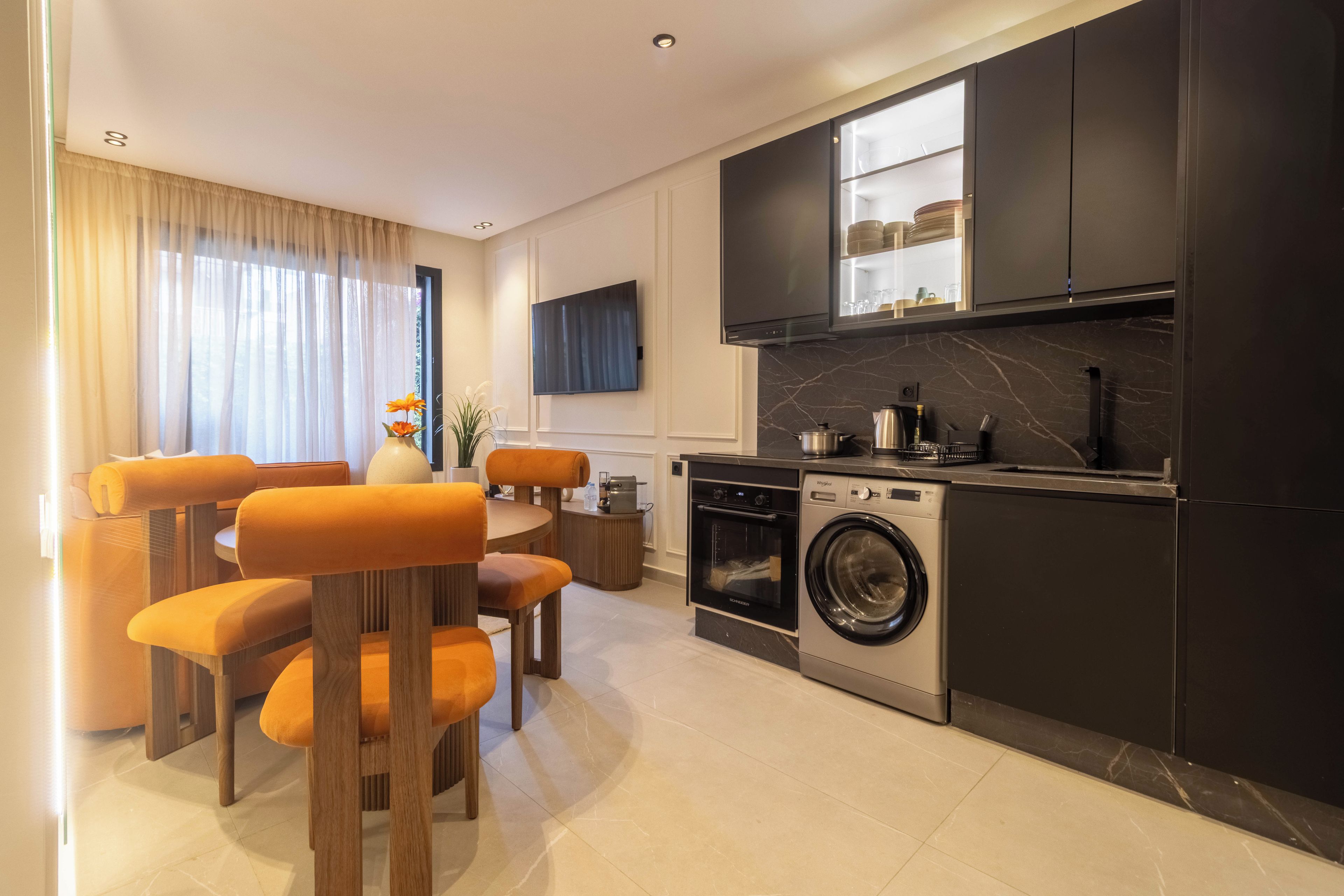 Eine moderne Küche mit Einbauofen, Mikrowelle und Waschmaschine, ein Essbereich mit orangefarbenen Stühlen und ein flachbildfernseher an der Wand.