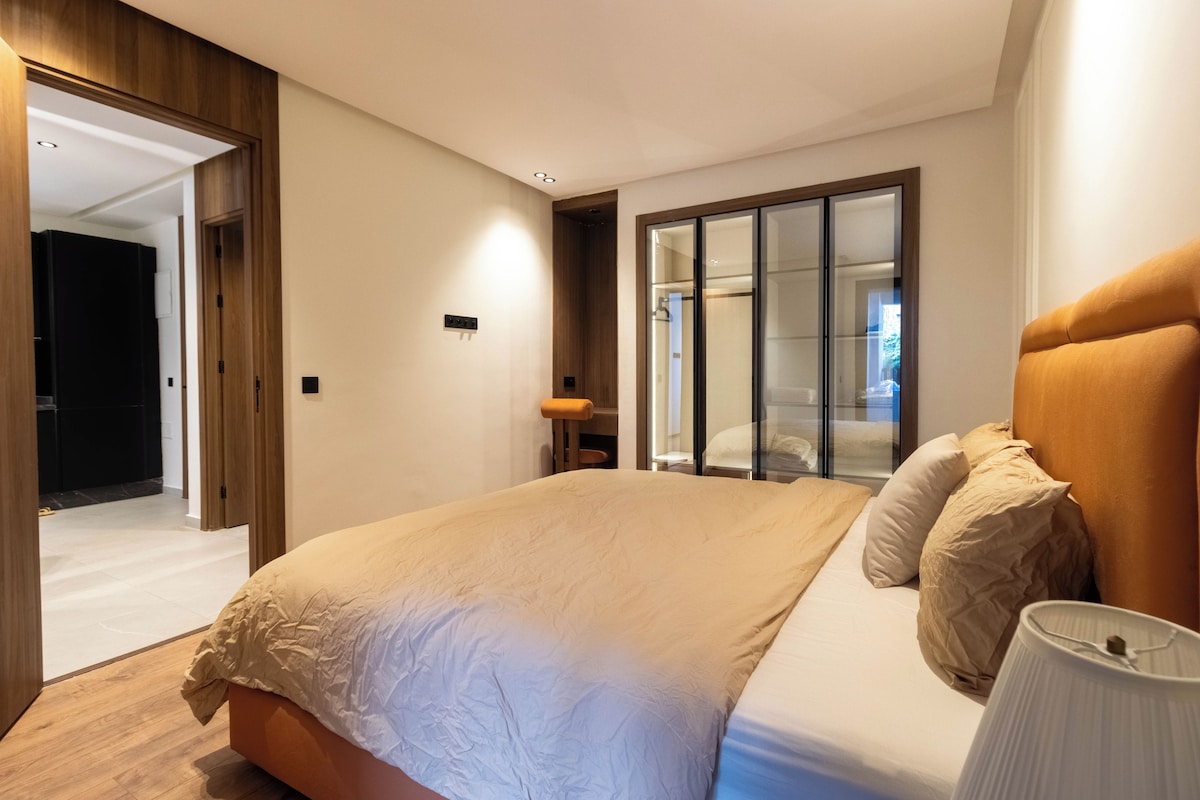 Ein modernes Schlafzimmer mit einem großen Bett, einem begehbaren Kleiderschrank und einem durch eine Glastür einsehbarer Badezimmer.