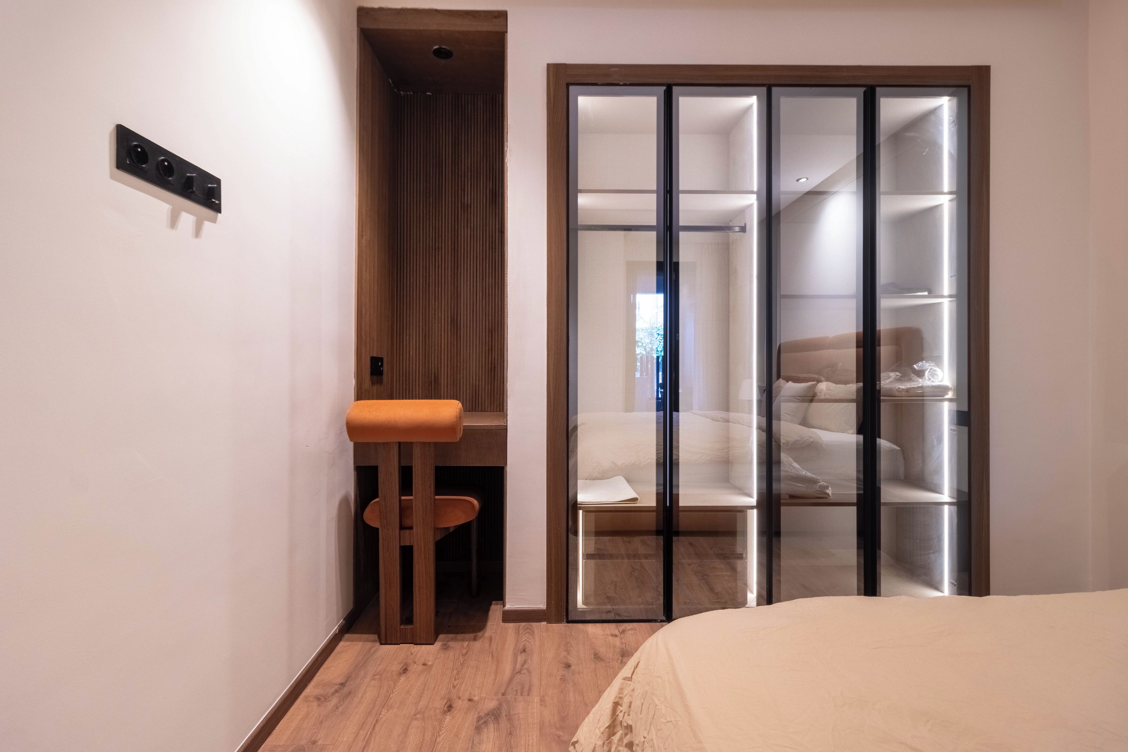 A bedroom with a bed, a wooden stool, a sliding glass closet, and a small window.