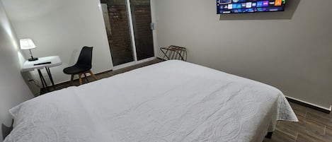 Un dormitorio con una cama grande, un escritorio con una lámpara, una silla, un ventanal corredero y un televisor montado que muestra varios canales.