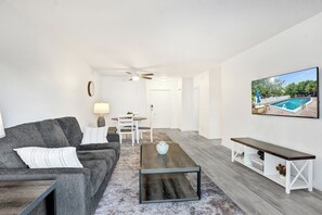 A living room with a grey sofa, a coffee table, a TV on the wall, and a view of a pool. - One block from the beach - Newly furnished 1 bedroom condo (Fort Lauderdale)