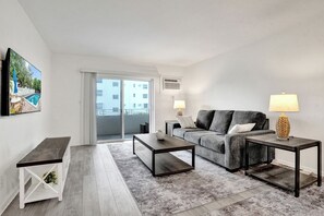 A modern living room with a grey sofa, two side tables, a coffee table, and a view of a pool and buildings through a sliding glass door. - One block from the beach - Newly furnished 1 bedroom condo (Fort Lauderdale)