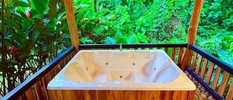 A hot tub under a wooden pergola with a stone floor, surrounded by lush greenery.