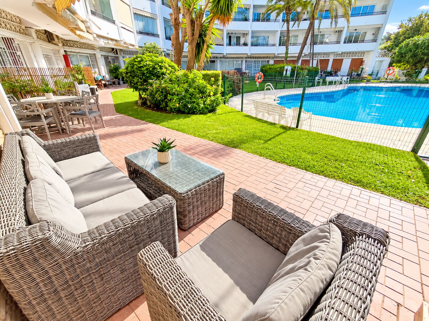 A poolside area with wicker furniture and a small table, surrounded by well-maintained grass and palm trees.