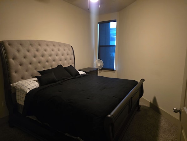 Ein Schlafzimmer mit einer gepolsterten Kopfteile, einem Bett mit schwarzer Bettwäsche, einem Nachttisch mit einem Ventilator und einem Fenster mit Blick.
