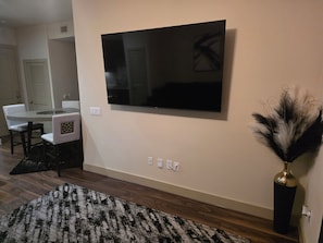 Ein modernes Wohnzimmer mit einem an der Wand befestigten Flachbildfernseher, einem Essbereich mit Tisch und Stühlen sowie einer dekorativen Vase mit Trockenpflanzen.
