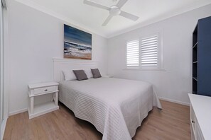 A bedroom with a bed, a nightstand, a ceiling fan, a window with blinds, and a painting on the wall. - Zilzie Beach House! (Zilzie)