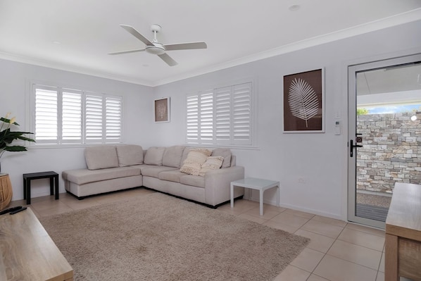 A living room with a beige sectional sofa, a small white side table, a ceiling fan, and a glass door leading to an outdoor area. - Zilzie Beach House! (Zilzie)