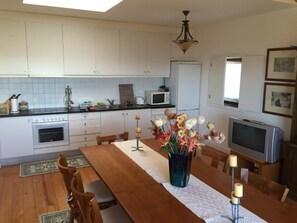 A kitchen with white cabinets, a microwave, a refrigerator, a wooden dining table with chairs, a vase with flowers, and a television.