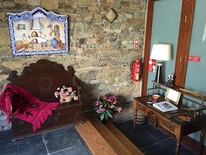 A room with a stone wall, a wooden bed with a red blanket, a wooden side table with a lamp, and a wall-mounted picture.