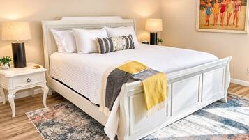 A bedroom with a white bed, two bedside tables, a nightstand, a patterned rug, and a painting on the wall.