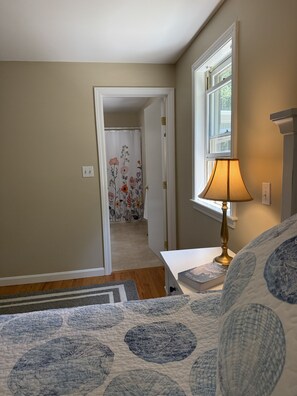 Un dormitorio con cama, una mesita de noche con lámpara, una ventana y una puerta que da a otra habitación.