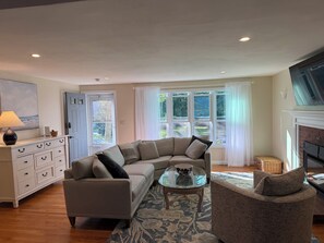 A living room with a sectional sofa, a glass coffee table, a side chair, a fireplace, and a large window with curtains.