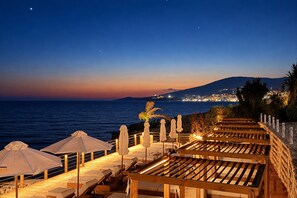 Ein Küstenort mit Sitzgelegenheiten auf einer Holzterrasse, Sonnenschirmen und Blick auf das Meer und die Stadtlichter.