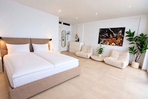 Ein modernes Schlafzimmer mit einem großen Bett, einer Sitzecke mit Sofas, einem kleinen Tisch und einer Topfpflanze.