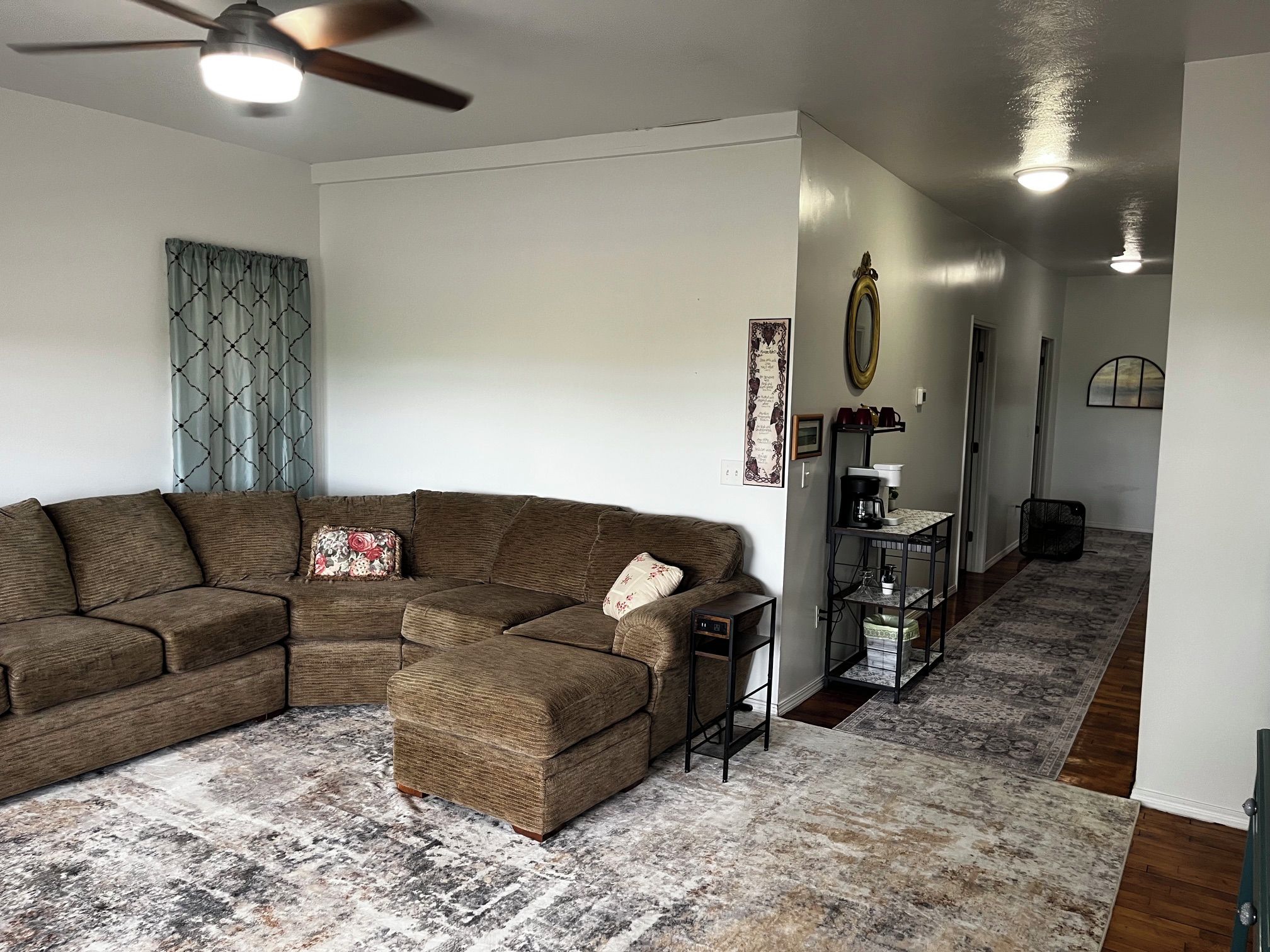 A living room with a sectional sofa, a coffee table, a patterned rug, a ceiling fan, and a hallway with a mirror and a coffee station.