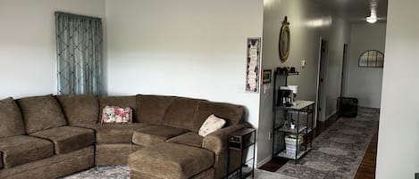 A living room with a sectional sofa, a coffee table, a patterned rug, a ceiling fan, and a hallway with a mirror and a coffee station.