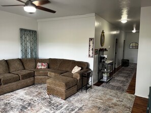 A living room with a sectional sofa, a coffee table, a patterned rug, a ceiling fan, and a hallway with a mirror and a coffee station. - CatalpaCorner 2-Bed Sit & Escape/Turnkey/Mid-term/Monthly/Weekly (Springfield)