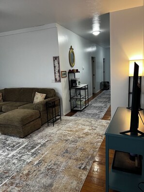 A living room with a sofa, a coffee table, a TV on a stand, a patterned rug, and a hallway with a door. - CatalpaCorner 2-Bed Sit & Escape/Turnkey/Mid-term/Monthly/Weekly (Springfield)