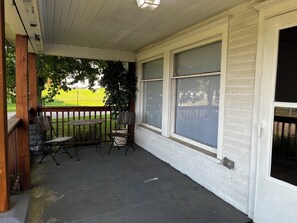 A covered porch with a table and chairs, a view of a green field, and a white building with large windows. - CatalpaCorner 2-Bed Sit & Escape/Turnkey/Mid-term/Monthly/Weekly (Springfield)