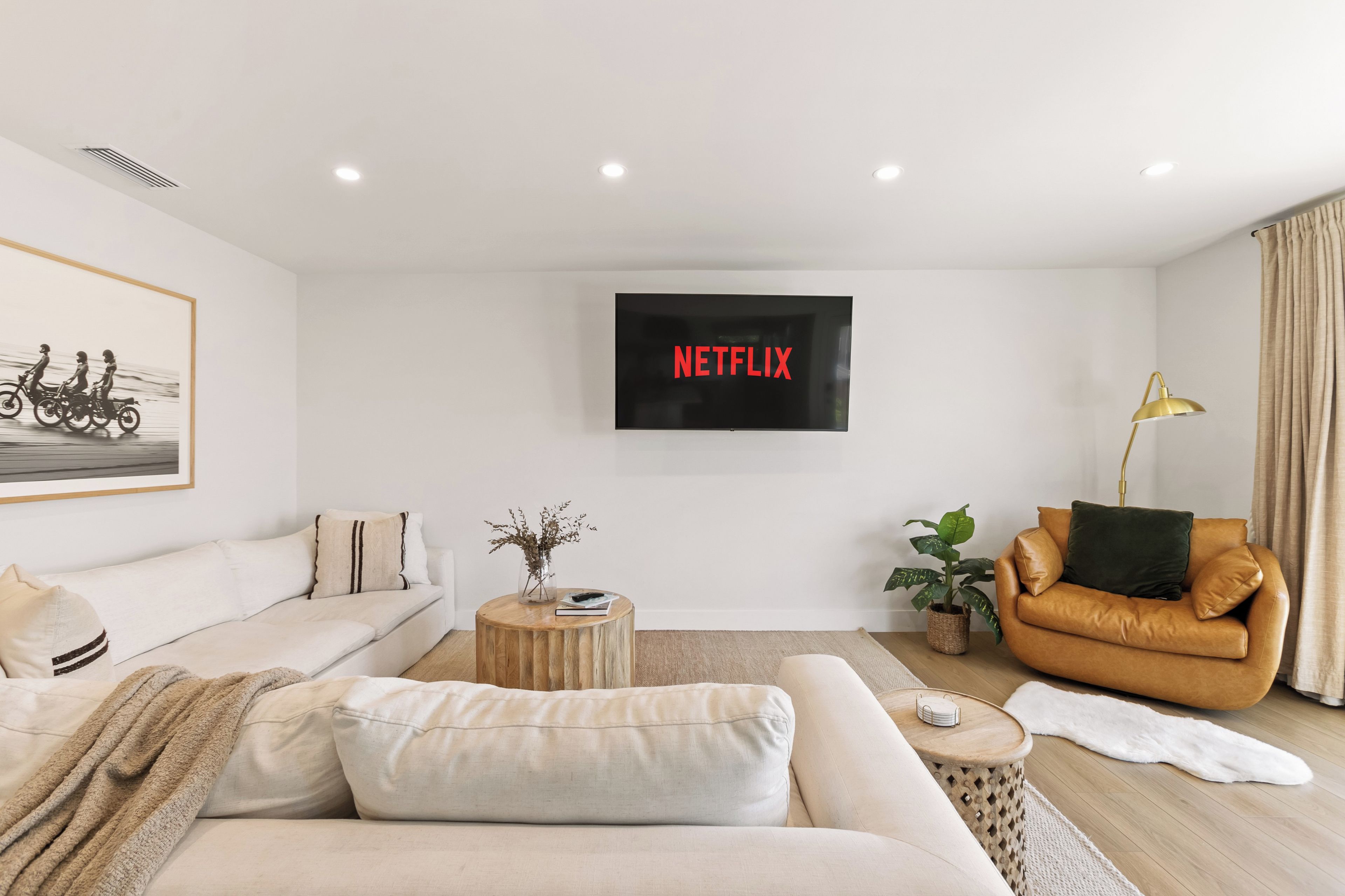 A modern living room with a sofa, a coffee table, a TV mounted on the wall, a brown leather armchair, and a potted plant.