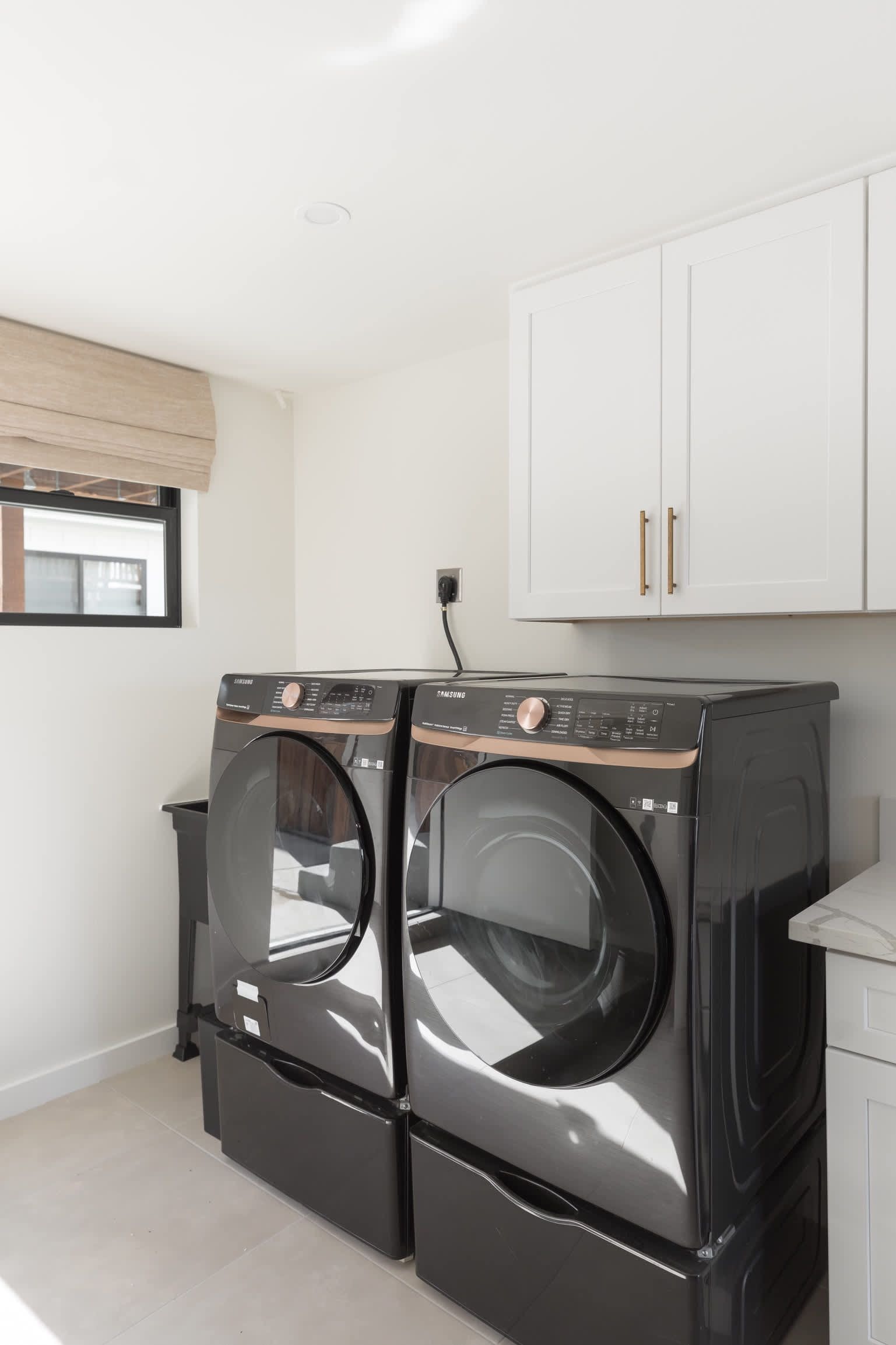 A laundry room with two black front-loading washing machines, a white cabinet above, and a window with blinds.